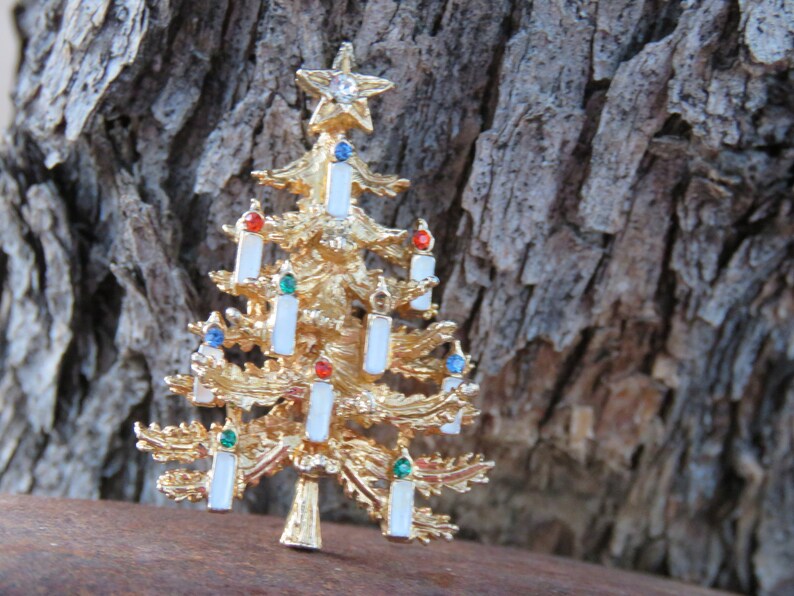 May include: A gold-toned Christmas tree brooch with white candle accents and colorful gems. The tree is topped with a star and has a textured surface. The brooch is approximately 2 inches tall.