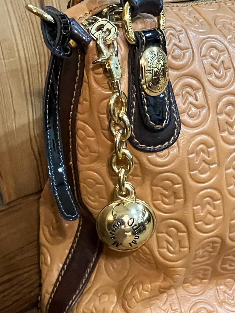 Pode incluir: Uma bolsa de m&atilde;o de couro bege com uma corrente dourada e um charme dourado que diz "Occhiali Made in Italy".