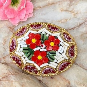 May include: A red, white, and green mosaic brooch with a floral design. The brooch is oval shaped and has a gold border.