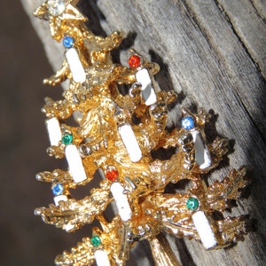May include: A gold-toned Christmas tree brooch with white enamel candle accents and colorful gemstone embellishments. The brooch features a star at the top with a clear rhinestone. A festive holiday accessory.