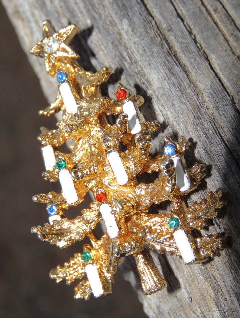 May include: A gold-toned Christmas tree brooch with white enamel candle accents and colorful gemstone embellishments. The brooch features a star at the top with a clear rhinestone. A festive holiday accessory.