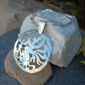 May include: A silver pendant necklace with a circular design featuring a detailed cutout of a figure with intricate floral patterns. The pendant is attached to a silver chain.