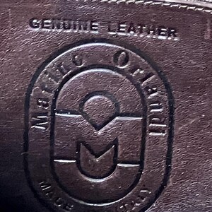 Pode incluir: Close-up de uma etiqueta de couro marrom com o texto "Genuine Leather" e um logotipo que diz "Made in Italy" com um "M" estilizado no centro.