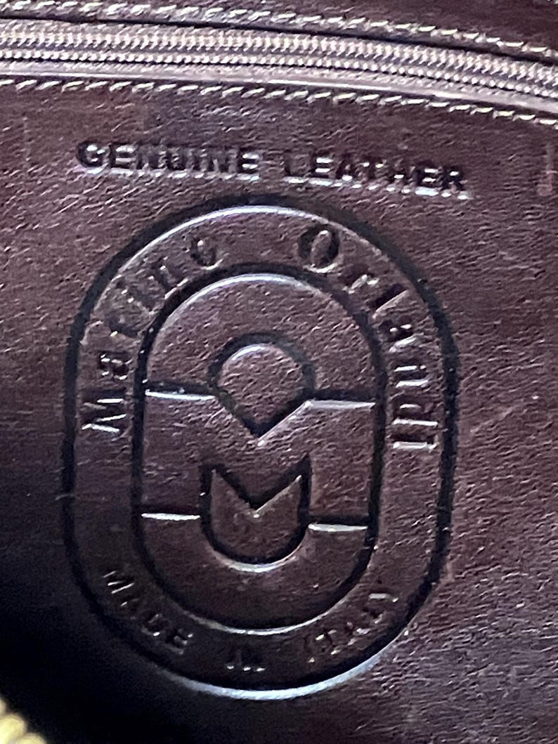 Pode incluir: Close-up de uma etiqueta de couro marrom com o texto "Genuine Leather" e um logotipo que diz "Made in Italy" com um "M" estilizado no centro.