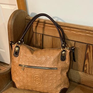 Pode incluir: Uma bolsa tote de couro marrom com um logotipo dourado em relevo e uma al&ccedil;a de couro marrom. A bolsa tem um fecho de z&iacute;per e um pequeno bolso na frente.