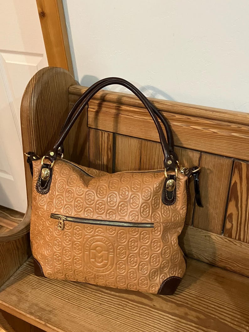 Pode incluir: Uma bolsa tote de couro marrom com um logotipo dourado em relevo e uma al&ccedil;a de couro marrom. A bolsa tem um fecho de z&iacute;per e um pequeno bolso na frente.