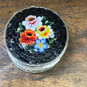 May include: A small, round trinket box with a mosaic floral design. The lid features a black background with colorful flowers in red, yellow, blue, and white. The box has a silver-colored metal frame and a textured edge.