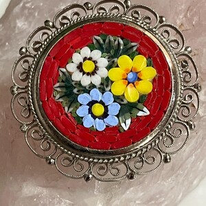 May include: A round, silver-toned brooch with a mosaic floral design. The center features a red background with three mosaic flowers in white, yellow, and blue. The brooch has a detailed, openwork border.