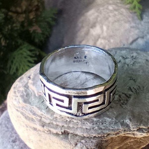 May include: A silver band ring with a Greek key design. The inside of the band is stamped "925 STERLING".