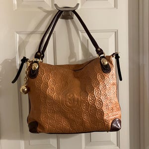 Pode incluir: Uma bolsa tote de couro marrom com um padr&atilde;o em relevo dourado e uma corrente dourada com um charme de bola. A bolsa tem al&ccedil;as de couro marrom e um z&iacute;per dourado.