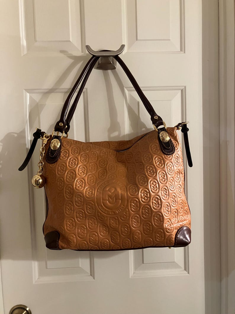 Pode incluir: Uma bolsa tote de couro marrom com um padr&atilde;o em relevo dourado e uma corrente dourada com um charme de bola. A bolsa tem al&ccedil;as de couro marrom e um z&iacute;per dourado.
