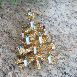 May include: Gold-toned Christmas tree brooch with a star on top. The tree features white candle accents with red, green, and blue gems. A festive holiday accessory.
