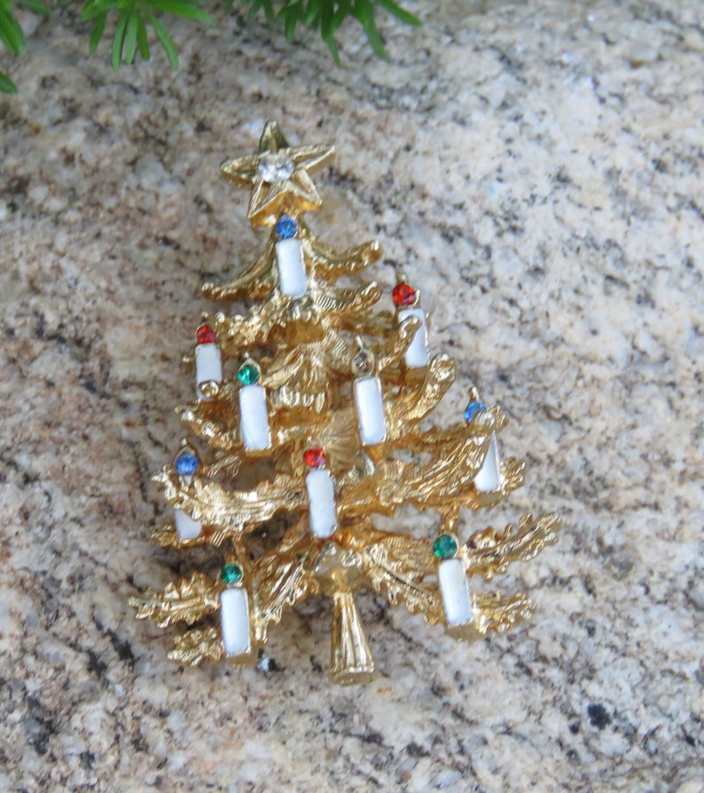 May include: Gold-toned Christmas tree brooch with a star on top. The tree features white candle accents with red, green, and blue gems. A festive holiday accessory.