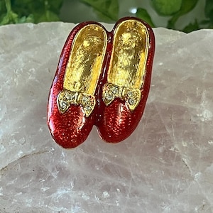 May include: A pair of red enamel shoes with gold interiors and rhinestone bows. The shoes are a miniature decorative item, possibly a brooch or pendant. The red color is vibrant and glossy, and the gold interior adds a touch of elegance.