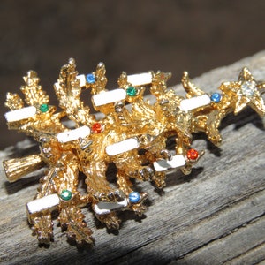 May include: A gold-toned Christmas tree brooch with white enamel accents and colorful gemstone embellishments. The brooch features a star at the top and is set against a rustic wooden surface.