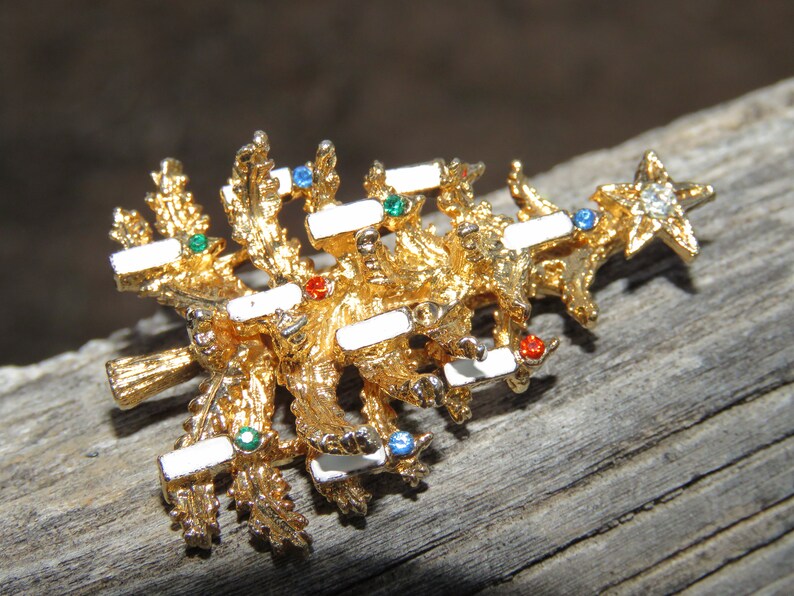 May include: A gold-toned Christmas tree brooch with white enamel accents and colorful gemstone embellishments. The brooch features a star at the top and is set against a rustic wooden surface.