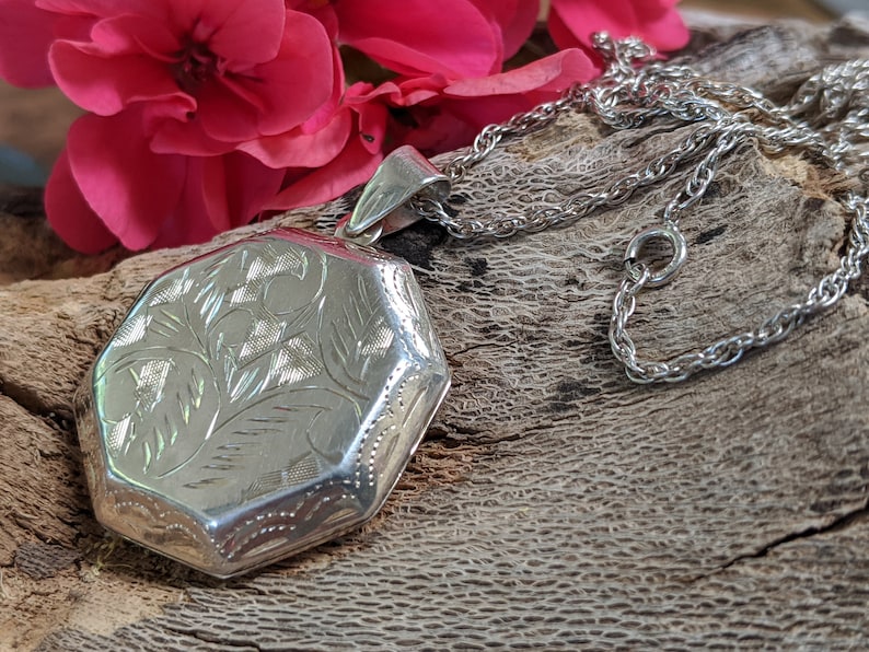 May include: A silver locket with an intricate floral design hangs from a silver chain. The locket is octagonal in shape and has a smooth, polished finish.