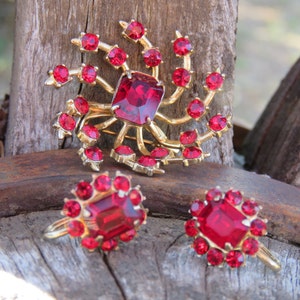 May include: A vintage jewelry set featuring a gold-toned brooch and matching earrings. The brooch has a large, square, red gemstone at its center, surrounded by smaller red stones. The earrings mirror the brooch design, with a square red stone and surrounding red accents.