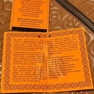 Pode incluir: Etiqueta laranja e branca com texto em ingl&ecirc;s, franc&ecirc;s, alem&atilde;o, italiano, polon&ecirc;s e russo. O texto trata da autenticidade dos artigos de couro e da garantia de venda. A etiqueta &eacute; feita de papel grosso e tem uma superf&iacute;cie texturizada.