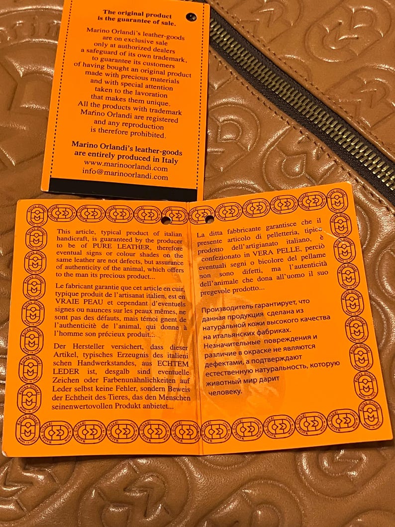 Pode incluir: Etiqueta laranja e branca com texto em ingl&ecirc;s, franc&ecirc;s, alem&atilde;o, italiano, polon&ecirc;s e russo. O texto trata da autenticidade dos artigos de couro e da garantia de venda. A etiqueta &eacute; feita de papel grosso e tem uma superf&iacute;cie texturizada.