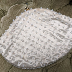 Puede incluir: Un bolso de mano de sat&eacute;n blanco con cierre dorado y dise&ntilde;o de cuentas. El bolso tiene forma de semic&iacute;rculo.