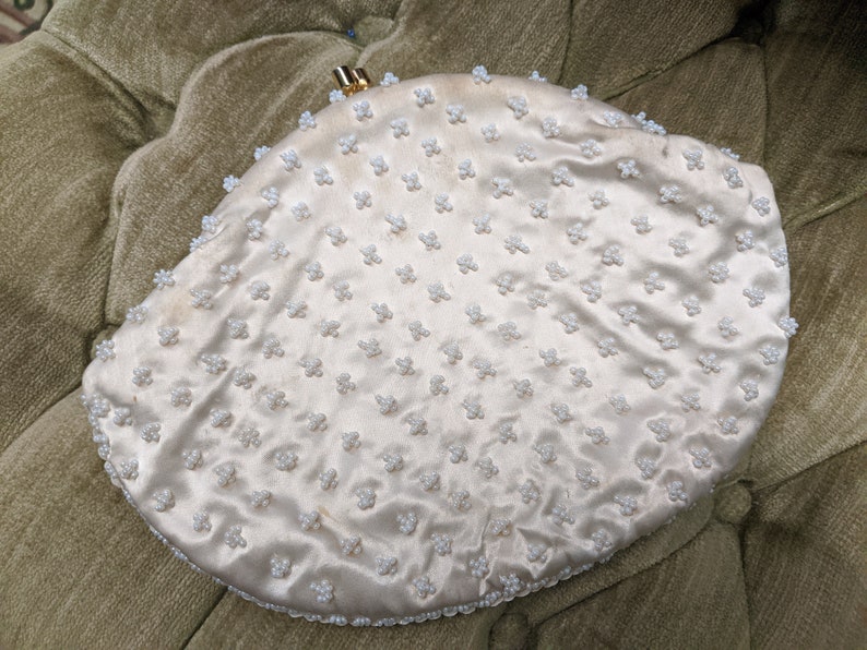 Puede incluir: Un bolso de mano de sat&eacute;n blanco con cierre dorado y dise&ntilde;o de cuentas. El bolso tiene forma de semic&iacute;rculo.