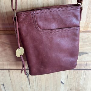 May include: A burgundy leather crossbody bag with a front pocket and adjustable strap. The bag features a gold-colored circular charm with a tassel. The bag is hanging against a wooden background.