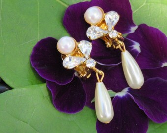 May include: A pair of gold tone clip-on earrings with a teardrop pearl dangle and crystal accents. The earrings are displayed on a purple flower.