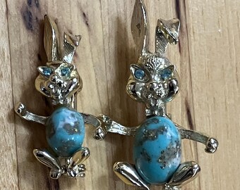 May include: Two gold-tone metal rabbit brooches of different sizes, each featuring a textured finish, blue rhinestone eyes, and a smooth, oval-shaped turquoise-colored stone with brown inclusions set into the center of the rabbit's body.