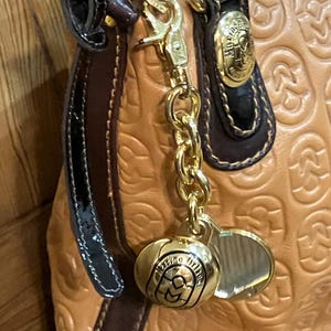 Pode incluir: Uma bolsa de m&atilde;o de couro bege com uma corrente dourada e um amuleto dourado que diz "Milano Italia". A bolsa tem uma al&ccedil;a de couro marrom e uma guarni&ccedil;&atilde;o de couro patenteado preto.