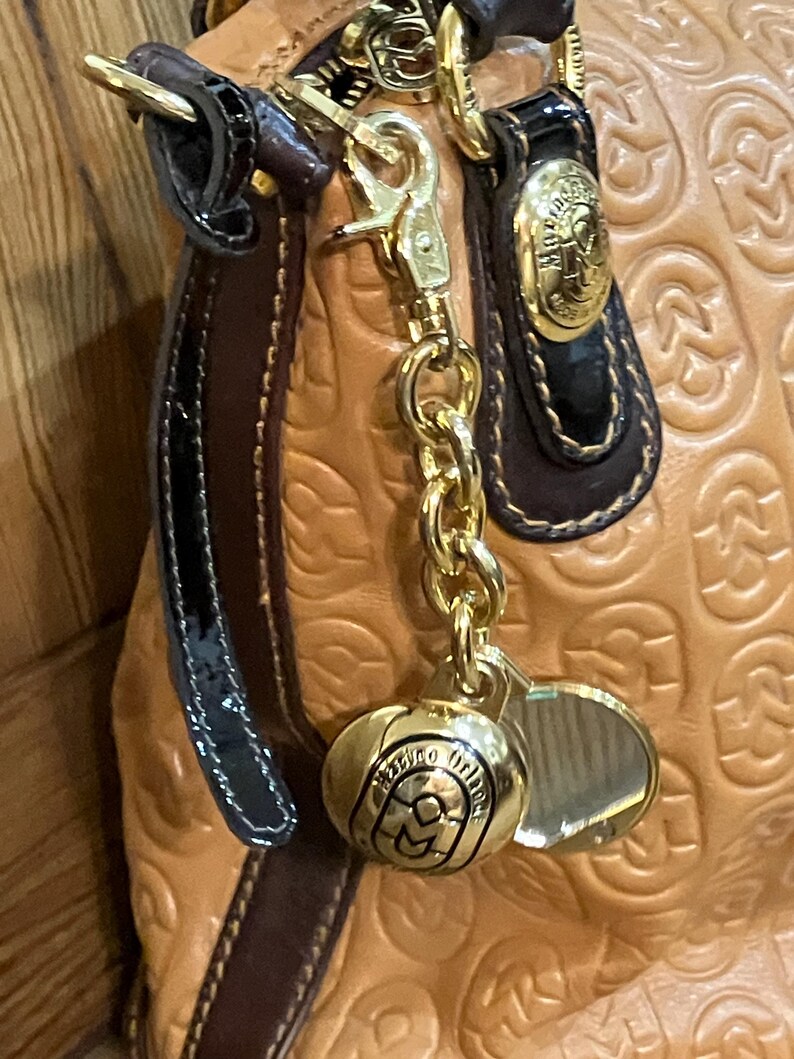 Pode incluir: Uma bolsa de m&atilde;o de couro bege com uma corrente dourada e um amuleto dourado que diz "Milano Italia". A bolsa tem uma al&ccedil;a de couro marrom e uma guarni&ccedil;&atilde;o de couro patenteado preto.