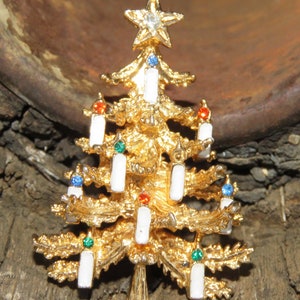 May include: Gold-toned Christmas tree brooch with a star on top and white candle accents. The brooch features colorful gems and is a festive holiday accessory.