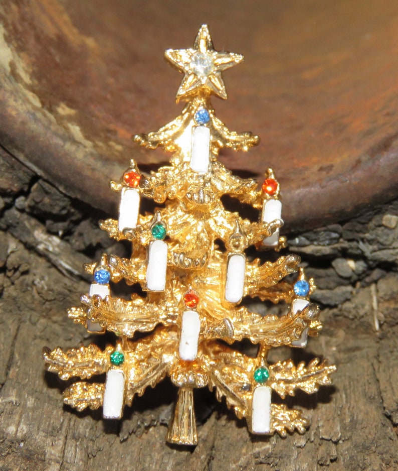 May include: Gold-toned Christmas tree brooch with a star on top and white candle accents. The brooch features colorful gems and is a festive holiday accessory.