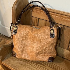 Pode incluir: Uma bolsa de m&atilde;o de couro marrom com al&ccedil;a de couro marrom e ferragens douradas. A bolsa tem um padr&atilde;o em relevo de c&iacute;rculos entrela&ccedil;ados e um logotipo que diz "Made in Italy".