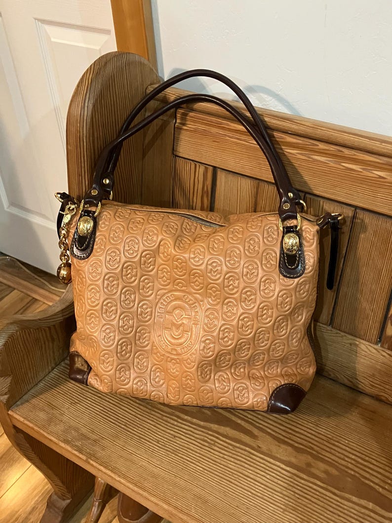 Pode incluir: Uma bolsa de m&atilde;o de couro marrom com al&ccedil;a de couro marrom e ferragens douradas. A bolsa tem um padr&atilde;o em relevo de c&iacute;rculos entrela&ccedil;ados e um logotipo que diz "Made in Italy".