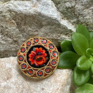 May include: A round, gold-toned brooch with a red and orange floral design in the center. The floral design is surrounded by a black border and a ring of gold stars.