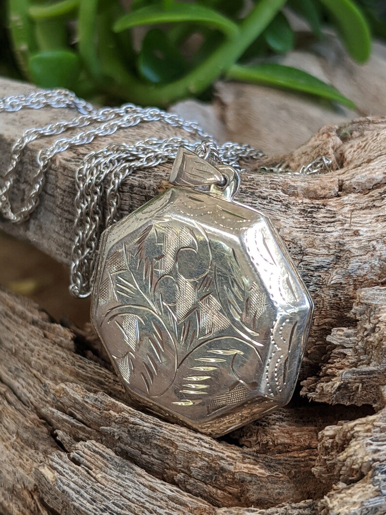 May include: A silver locket necklace with an octagonal pendant featuring an intricate floral design. The locket is on a silver chain.