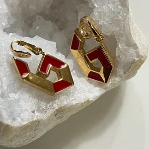 May include: A pair of gold-tone clip-on earrings featuring a geometric, open-frame design with vibrant red enamel accents. The earrings have a polished metallic finish and a secure clip mechanism on the back for attachment to the earlobe.
