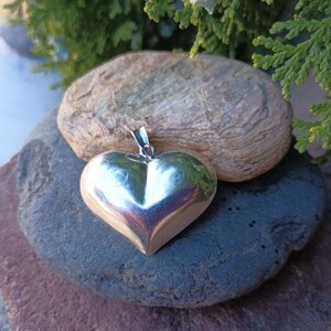 May include: A silver heart-shaped pendant with a smooth, shiny finish. The pendant is resting on a gray rock.