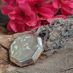 May include: A silver locket with an octagonal shape and an engraved floral design. The locket is attached to a silver chain.