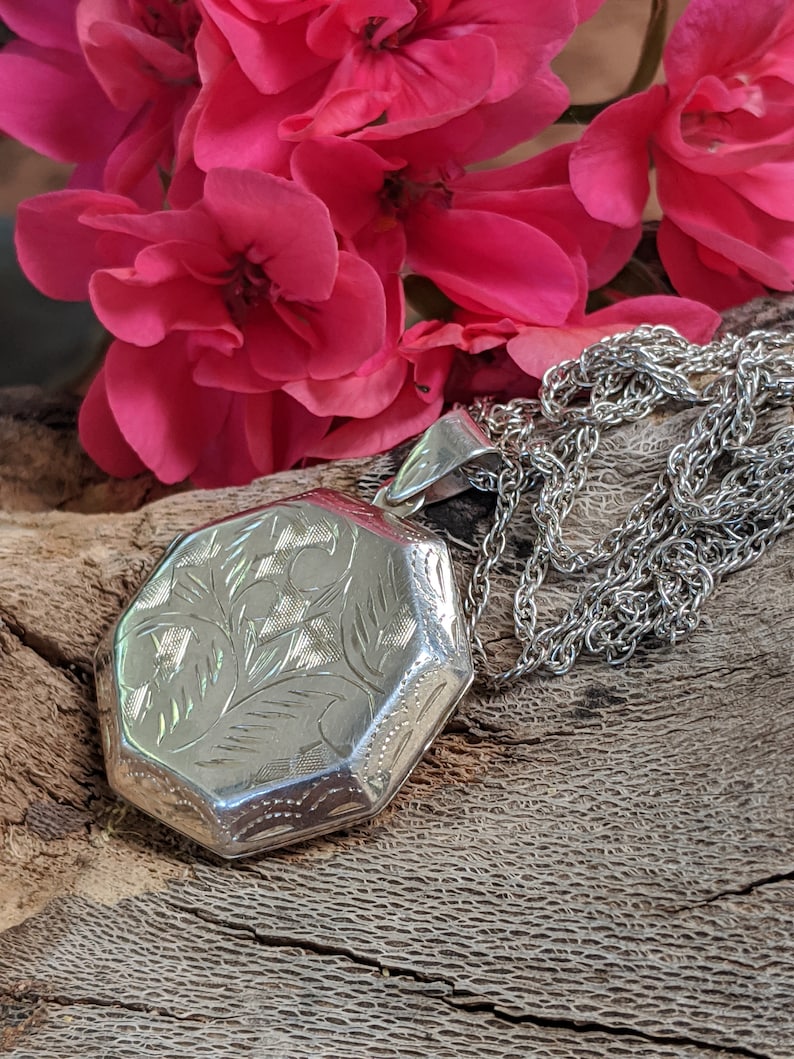 May include: A silver locket with an octagonal shape and an engraved floral design. The locket is attached to a silver chain.