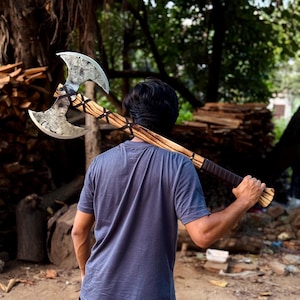 May include: A double-bit battle axe featuring a large, weathered metal head with two curved blades and a long wooden handle. The handle displays a distinct wood grain pattern and is wrapped with dark leather straps for a secure grip.