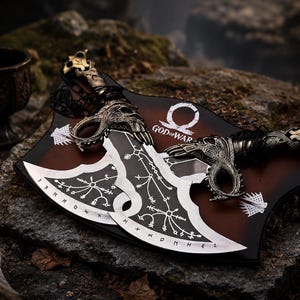 May include: Two silver-toned metal blades with intricate etched patterns and runic symbols, featuring ornate handles with skull motifs. The blades are mounted on a dark brown wooden plaque displaying the white God of War logo and text.
