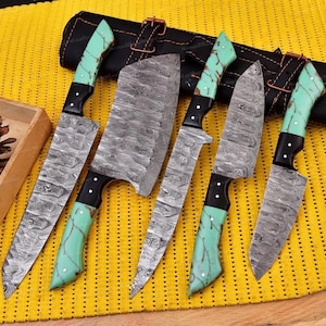 May include: A set of five kitchen knives featuring Damascus steel blades with a wavy pattern and handles made of black and mint green marbled material with silver rivets, arranged alongside a black leather knife roll.