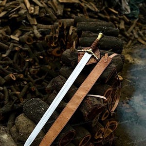 May include: A double-edged steel sword with a brown leather-wrapped grip and a brass pommel. The crossguard features a gold-colored metal design. A matching brown leather scabbard with a strap lies beside the blade on a pile of wood.