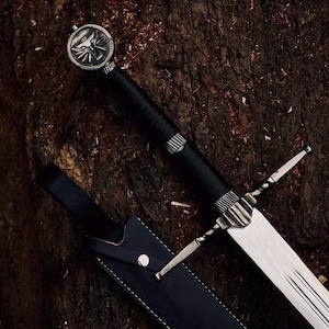 May include: A steel sword with a black leather-wrapped handle and a silver-toned pommel featuring a wolf head emblem. The crossguard is silver-toned with a twisted design. A black leather scabbard with white stitching and a silver snap button is included.
