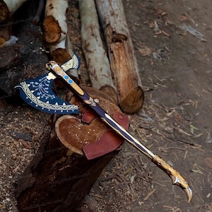 May include: A decorative battle axe featuring a dark blue blade with white etched Norse-inspired patterns and gold trim. The long wooden handle is accented with gold metal fittings, blue circular inlays, and a curved bird-shaped pommel.