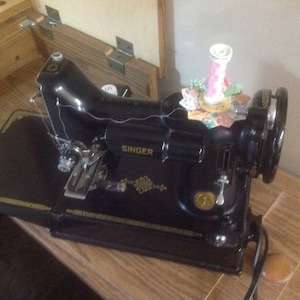 May include: A black Singer sewing machine with a gold decorative design. The machine has a white bobbin and a spool of thread on top.