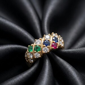 May include: A gold band ring featuring a multi-row design with round-cut gemstones. The ring is set with clear white diamonds, two green emeralds, two blue sapphires, and one red ruby, arranged in a diagonal pattern across the surface of the band.