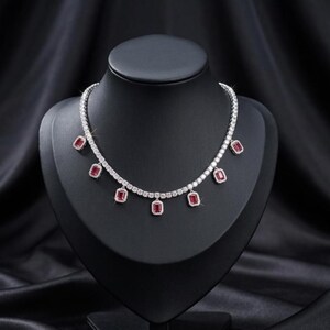 May include: A silver-toned tennis necklace featuring a continuous row of clear, round-cut stones. The necklace is accented with six evenly spaced, rectangular-cut red stones, each surrounded by a halo of small, clear, round-cut stones.